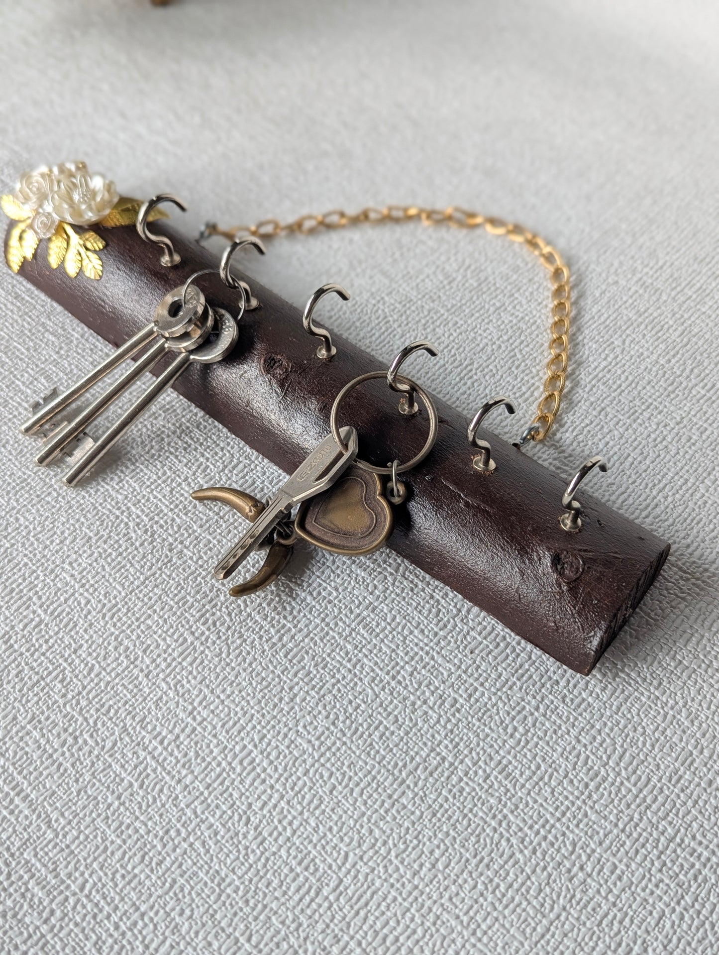 Handcrafted wooden key holder with floral accent, metal hooks, and gold chain, on textured surface